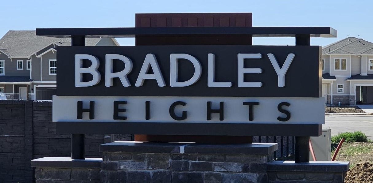 Blue skies with a modern monument sign that reads BRADLEY HEIGHTS with bricks on the bottom. There are two-story houses in the background.